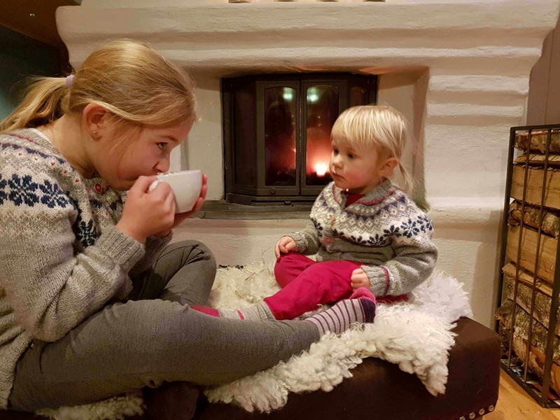Ei jente drikker kakao foran peisen mens lillesøster ser på